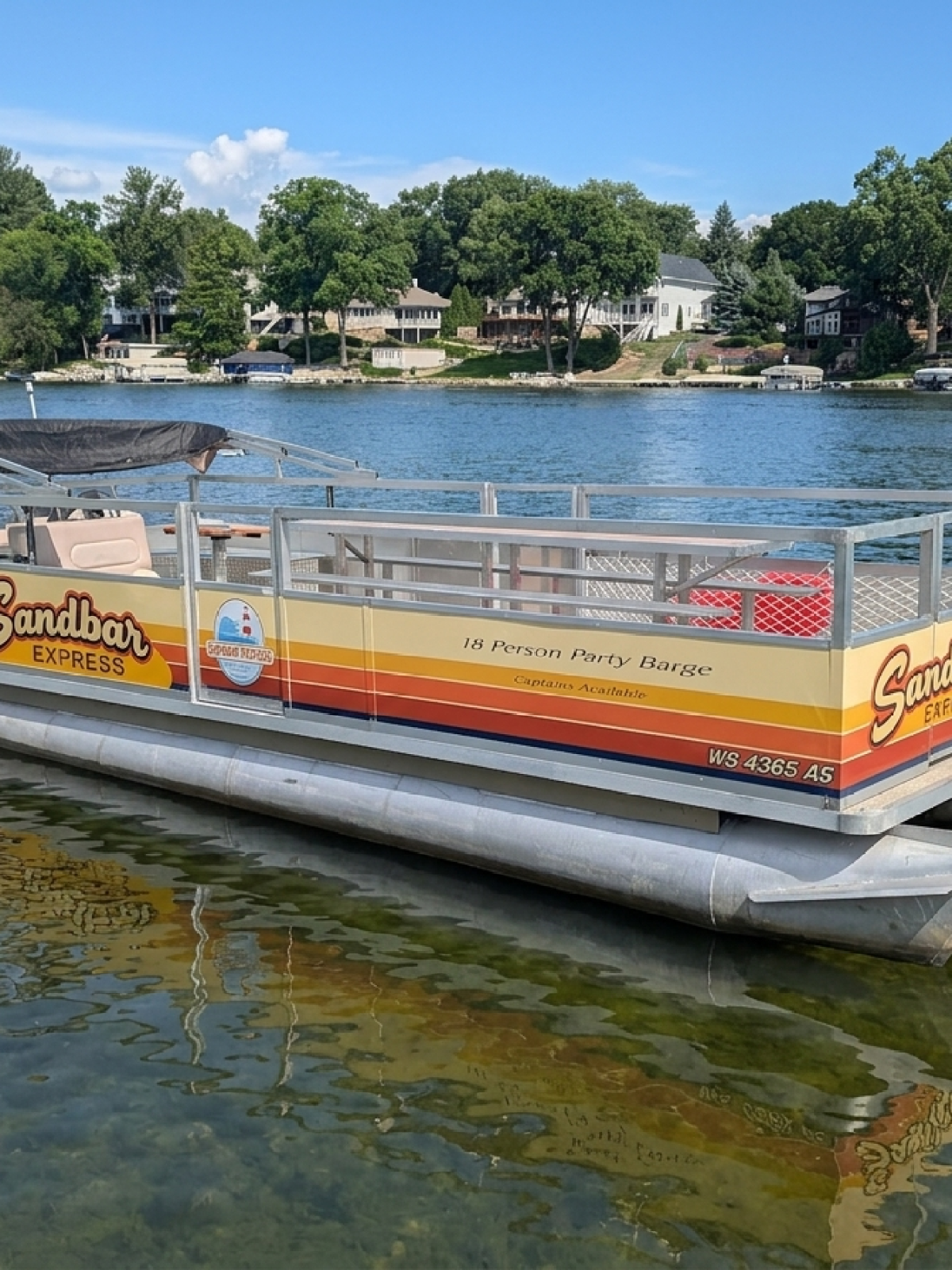 Pontoon boat named 'Sandbar Express' on a lake with trees and houses in the background.