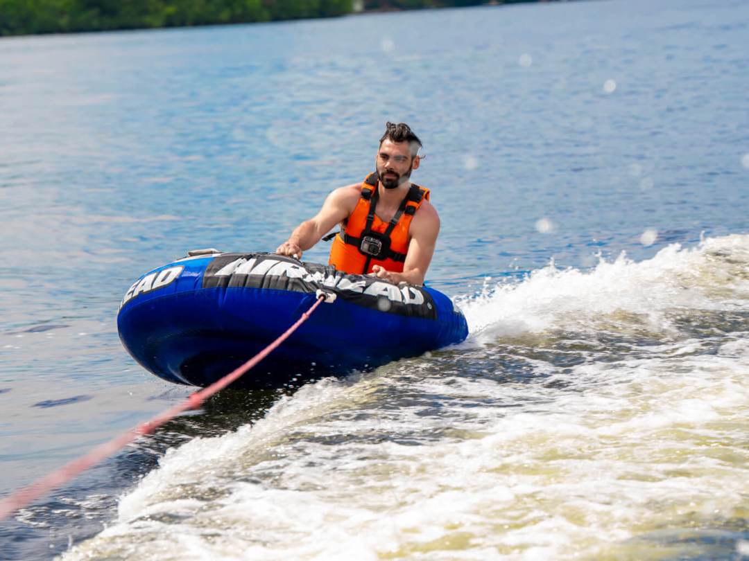 Person tubing on a lake wearing an orange life vest, being pulled by a boat.